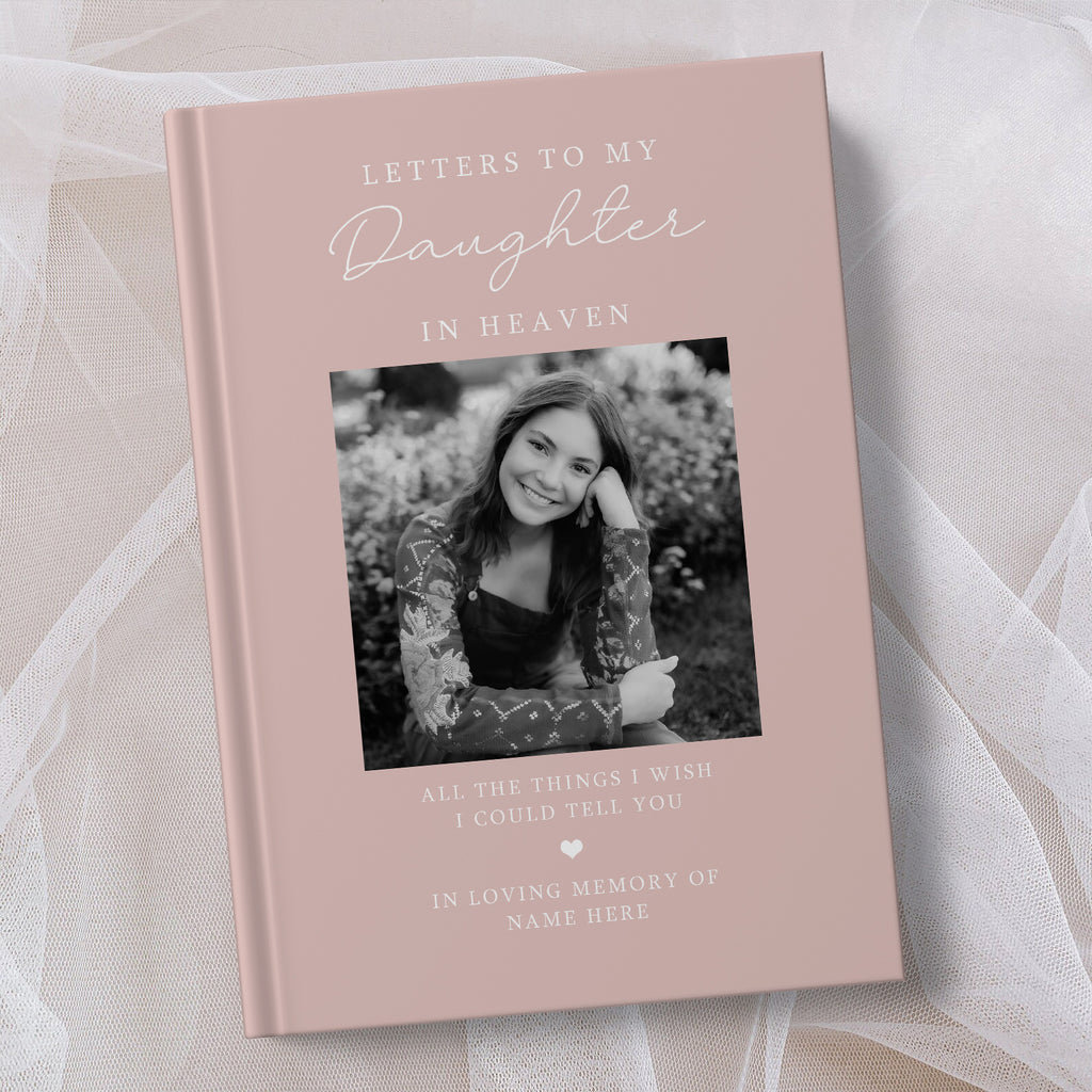 A personalised hardcover book titled 'Letters to My Daughter in Heaven' in a dusty pink colour, featuring a black and white photo of a young woman on the cover, displayed on a soft white fabric background.