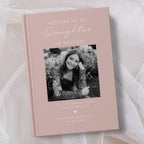 A personalised hardcover book titled 'Letters to My Daughter in Heaven' in a dusty pink colour, featuring a black and white photo of a young woman on the cover, displayed on a soft white fabric background.