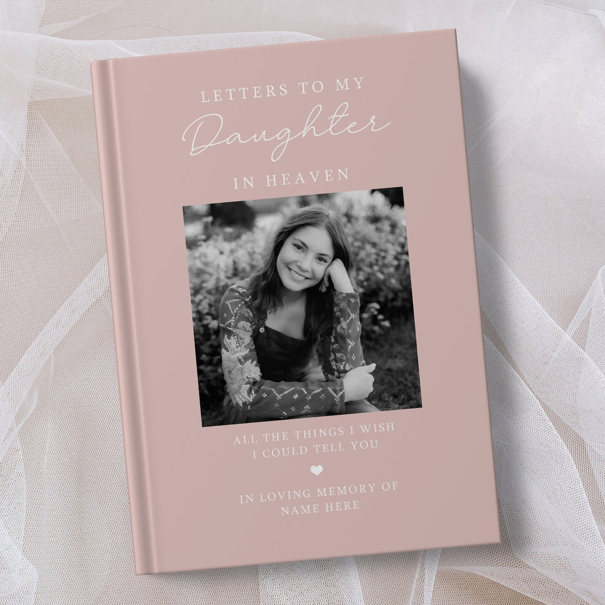 A personalised hardcover book titled 'Letters to My Daughter in Heaven' in a dusty pink colour, featuring a black and white photo of a young woman on the cover, displayed on a soft white fabric background.