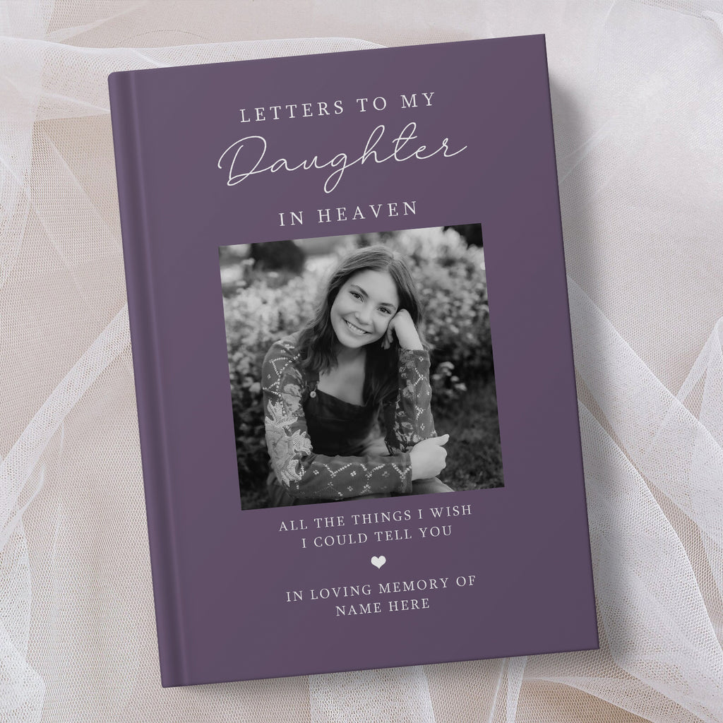 A personalised hardcover book with a muted purple cover, featuring a black and white photo of a young woman and placeholder text for a memorial letter to her in heaven, styled on a soft beige fabric background.