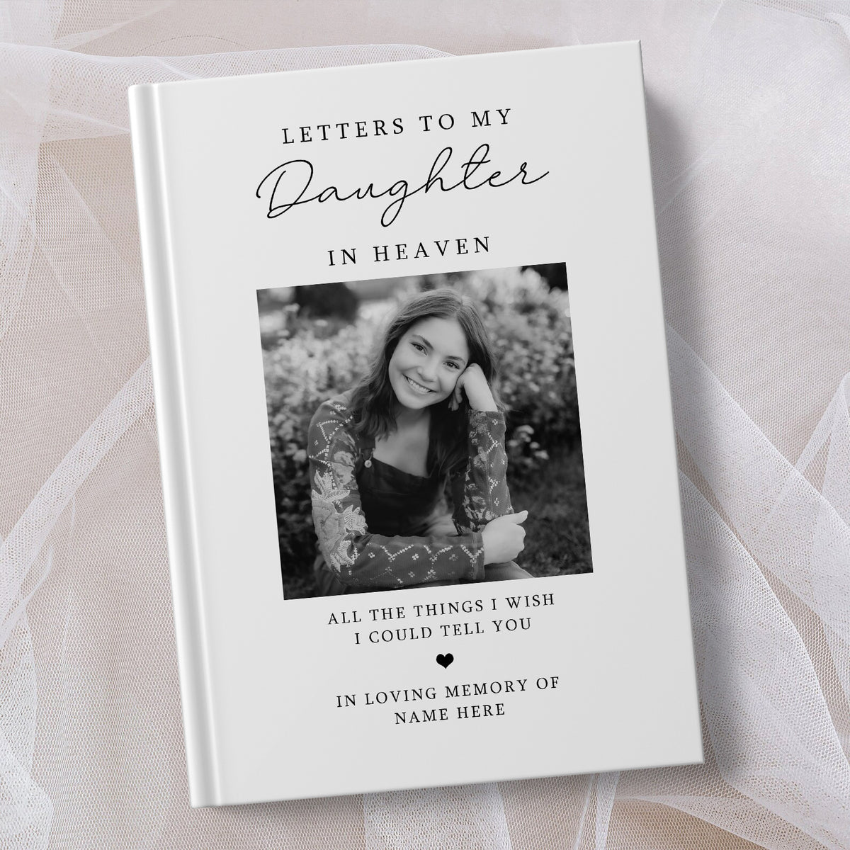 A personalised hardcover book titled 'Letters to My Daughter in Heaven' with a black and white photo of a woman on the cover, styled on a soft beige fabric background.