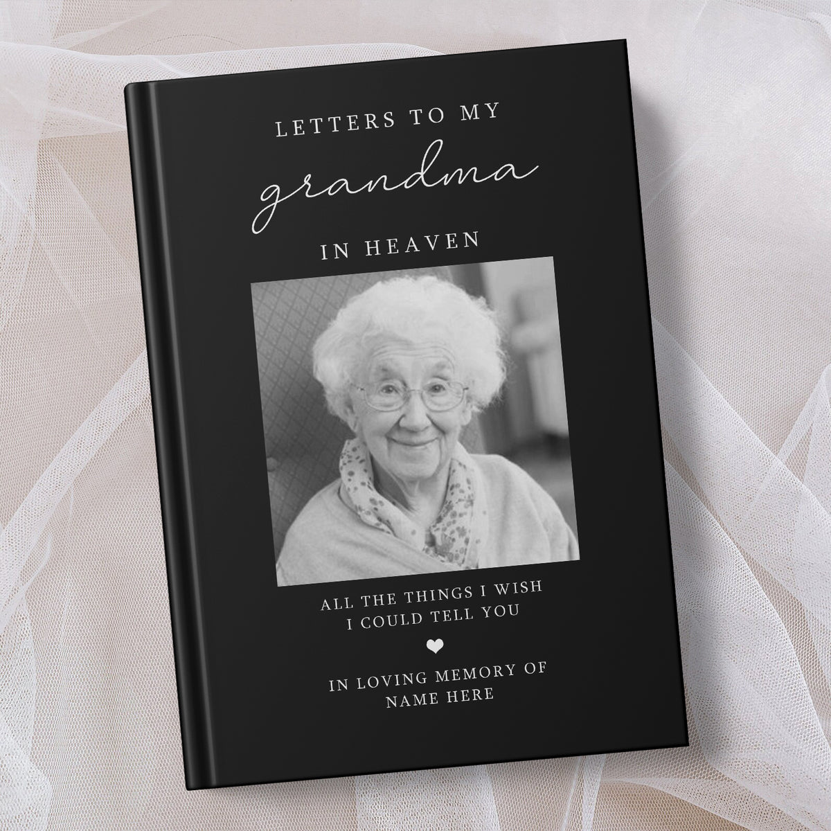 A personalised hardcover memorial book titled 'Letters to My Grandma in Heaven' with a black cover, white text, and a black and white photo of an elderly woman. The book is photographed on a soft white fabric background.