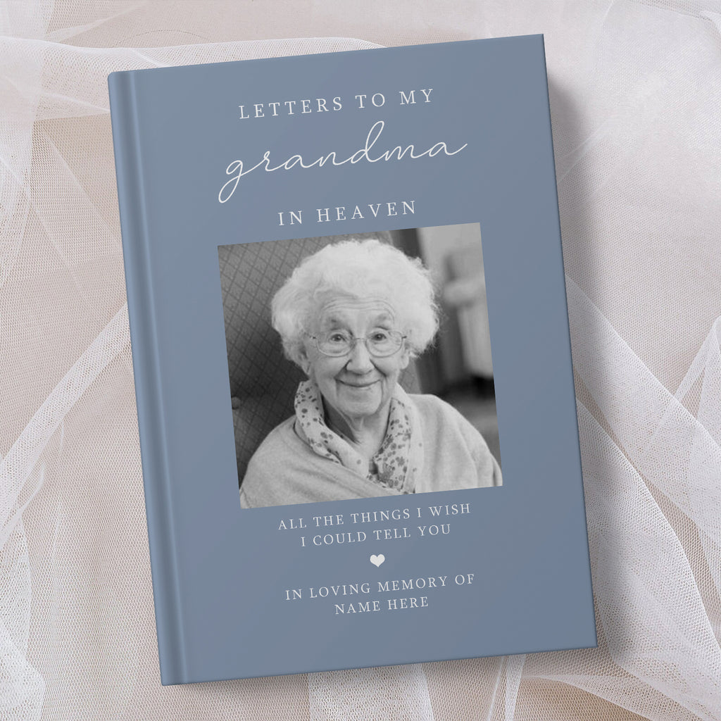 A personalised hardcover book titled 'Letters to My Grandma in Heaven' with a slate blue cover, a black and white photo of an elderly woman, and placeholder text for a memory message, styled on a soft beige fabric background.
