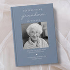 A personalised hardcover book titled 'Letters to My Grandma in Heaven' with a slate blue cover, a black and white photo of an elderly woman, and placeholder text for a memory message, styled on a soft beige fabric background.