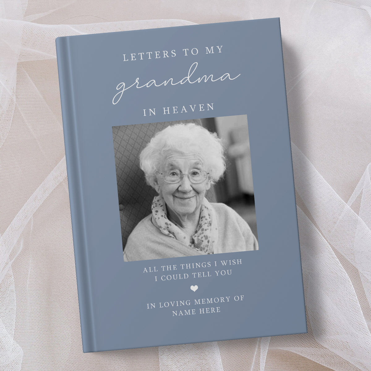 A personalised hardcover book titled 'Letters to My Grandma in Heaven' with a slate blue cover, a black and white photo of an elderly woman, and placeholder text for a memory message, styled on a soft beige fabric background.