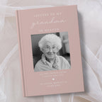 A personalised hardcover book titled 'Letters to My Grandma in Heaven' with a dusty pink cover, featuring a black and white photo of an elderly woman and placeholder text, displayed on a soft white fabric background.