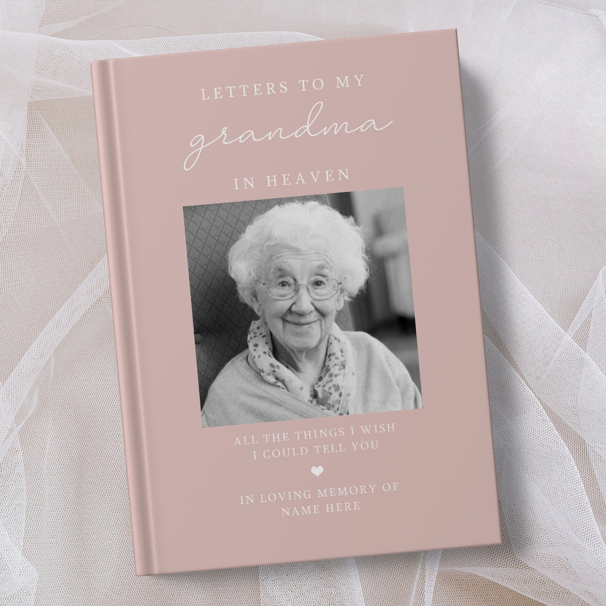 A personalised hardcover book titled 'Letters to My Grandma in Heaven' with a dusty pink cover, featuring a black and white photo of an elderly woman and placeholder text, displayed on a soft white fabric background.