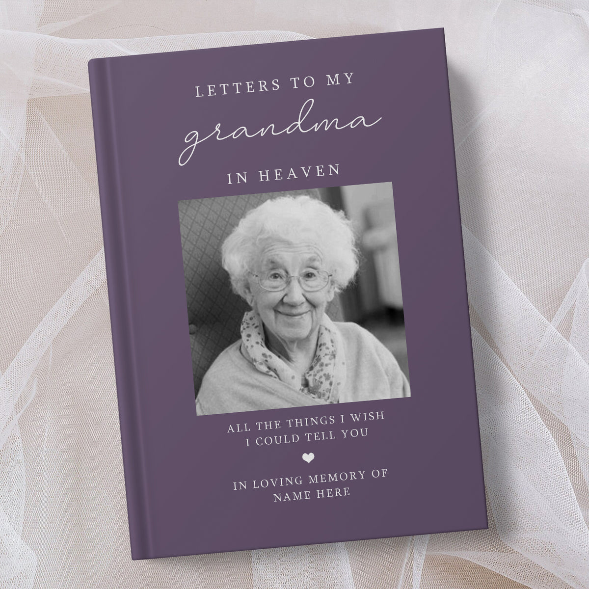 A personalised hardcover memorial book titled 'Letters to My Grandma in Heaven' with a purple cover, a black and white photo of an elderly woman, and placeholder text for personalisation, styled on soft white tulle fabric.