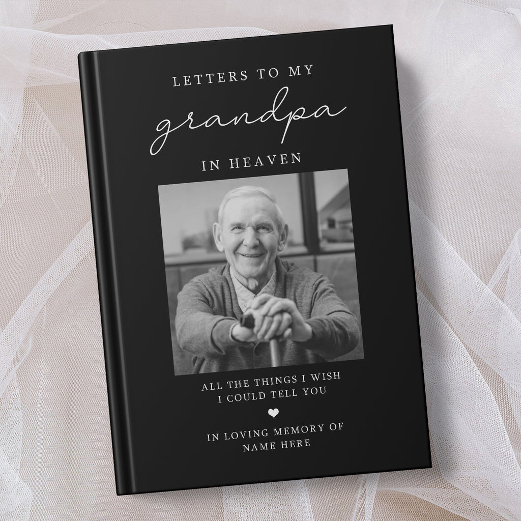 A personalised hardcover book titled 'Letters to My Grandpa in Heaven' with a black cover, white text, and a placeholder photo of an elderly man. The book is displayed on a soft, draped white fabric background.