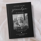 A personalised hardcover book titled 'Letters to My Grandpa in Heaven' with a black cover, white text, and a placeholder photo of an elderly man. The book is displayed on a soft, draped white fabric background.