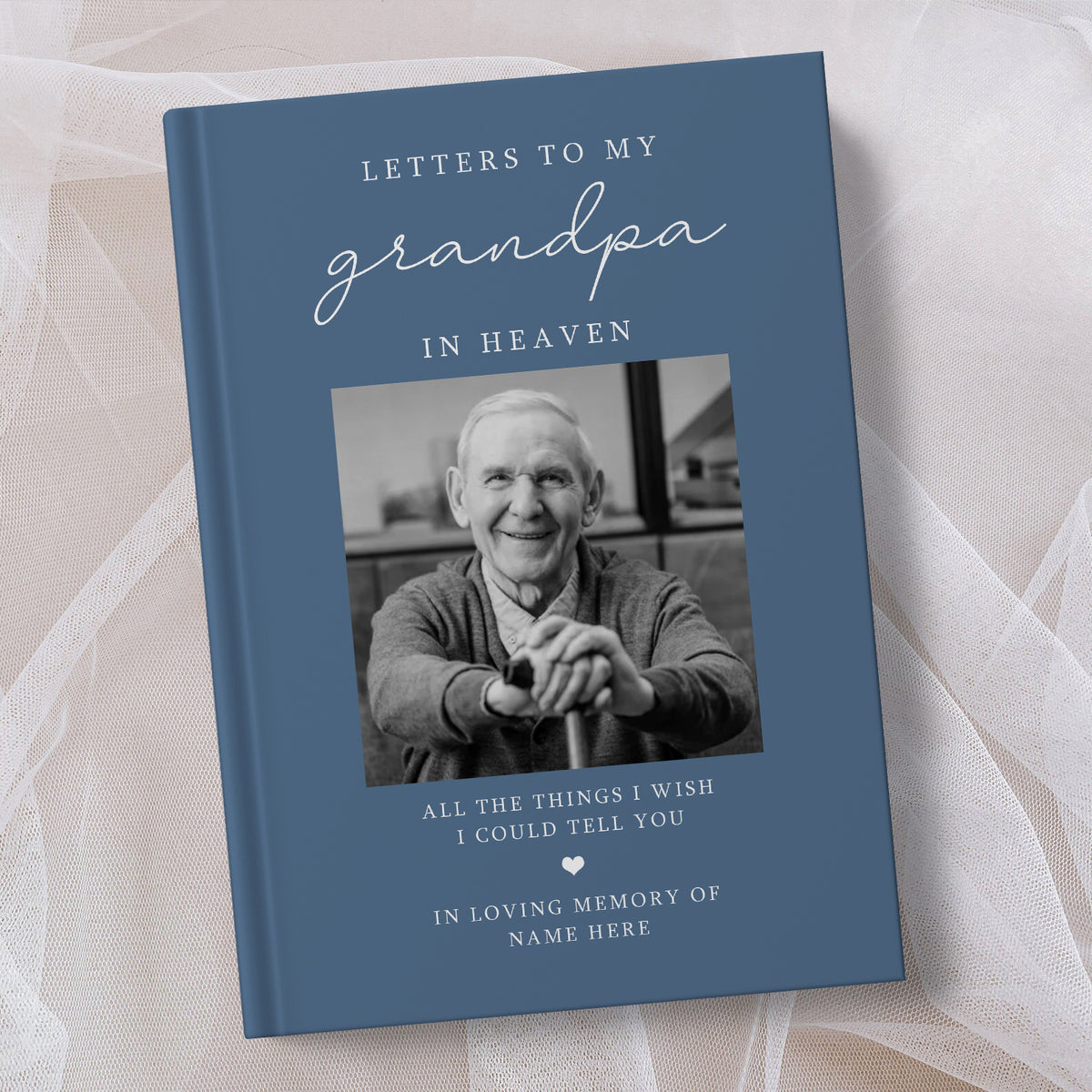 A personalised hardcover book titled 'Letters to My Grandpa in Heaven' with a slate blue cover, a black and white portrait photo of an elderly man, and white text, styled on a soft beige fabric background.