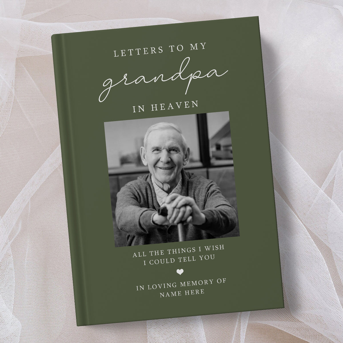 A personalised hardcover book titled 'Letters to My Grandpa in Heaven' with an olive green cover, featuring a placeholder black and white photo of an elderly man. The book is displayed on a soft, draped white fabric background.