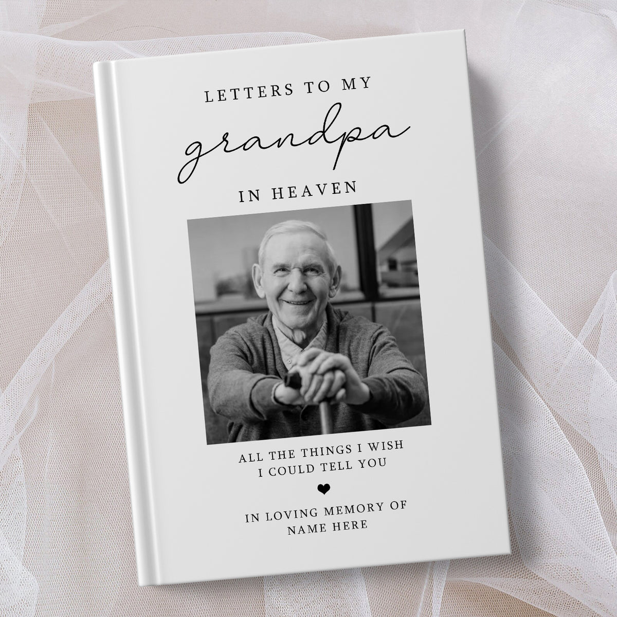 A personalised hardcover book titled 'Letters to My Grandpa in Heaven' with a black and white portrait photo of an elderly man on the cover, styled on a soft white fabric background.