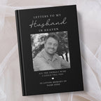 A personalised hardcover book titled 'Letters to My Husband in Heaven' with a black cover, white text, and a black and white photo of a smiling man. The book is displayed on a soft beige fabric background.