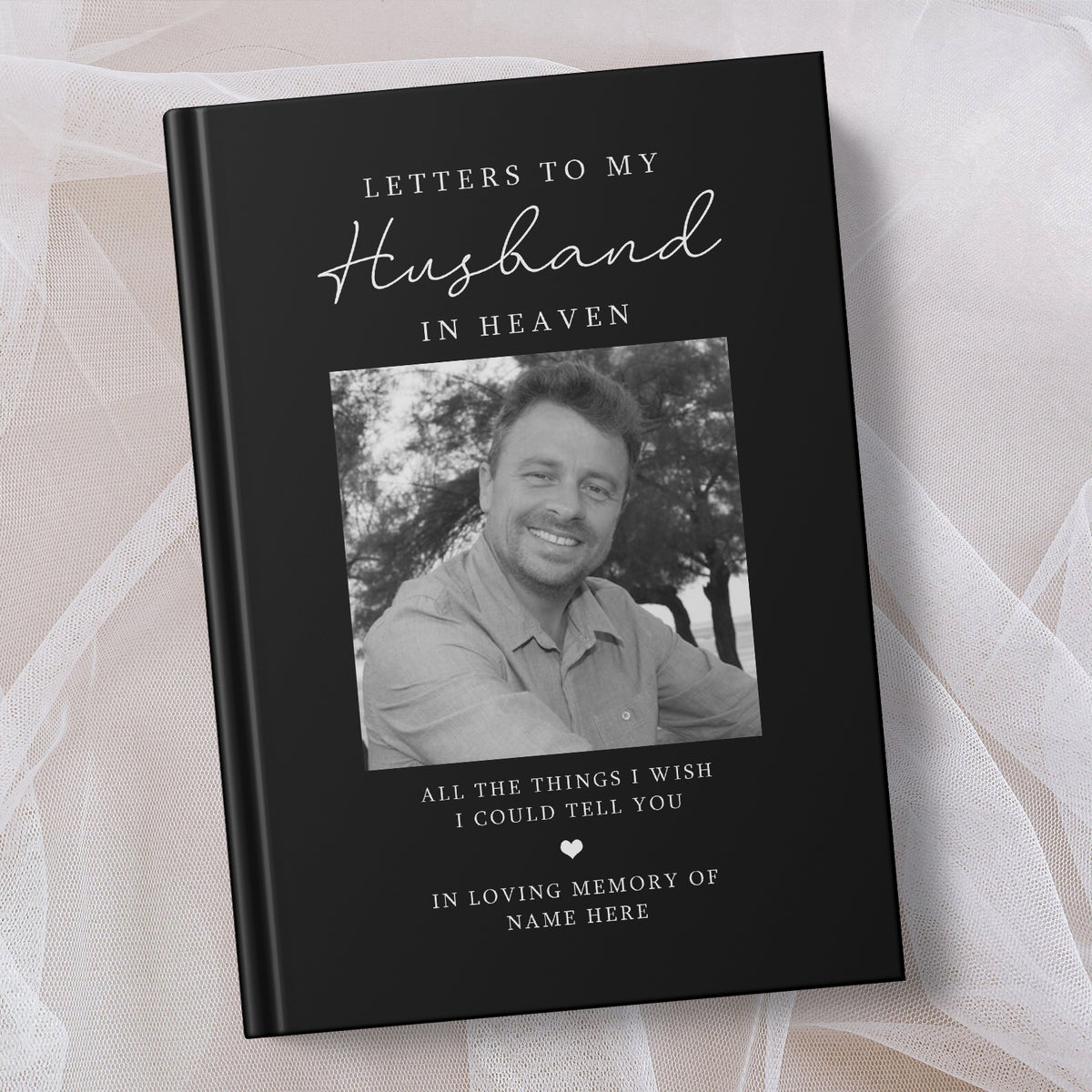 A personalised hardcover book titled 'Letters to My Husband in Heaven' with a black cover, white text, and a black and white photo of a smiling man. The book is displayed on a soft beige fabric background.