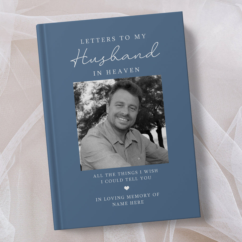 A personalised hardcover book titled 'Letters to My Husband in Heaven' in slate blue, featuring a black and white photo of a smiling man on the cover, displayed on a soft white fabric background.