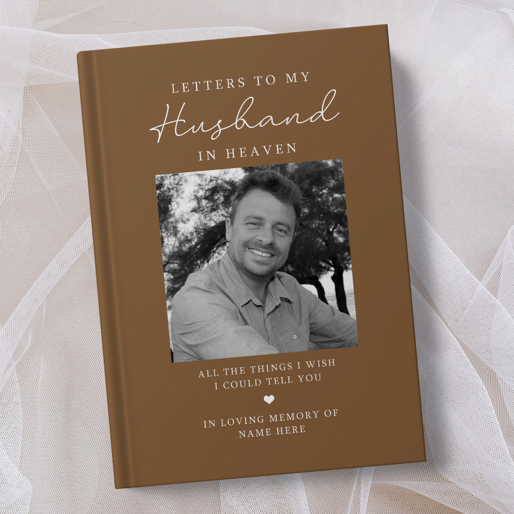 A personalised hardcover book titled 'Letters to My Husband in Heaven' with a brown cover, white elegant text, and a black and white photo of a smiling man. The book is displayed on a soft, draped white fabric background.