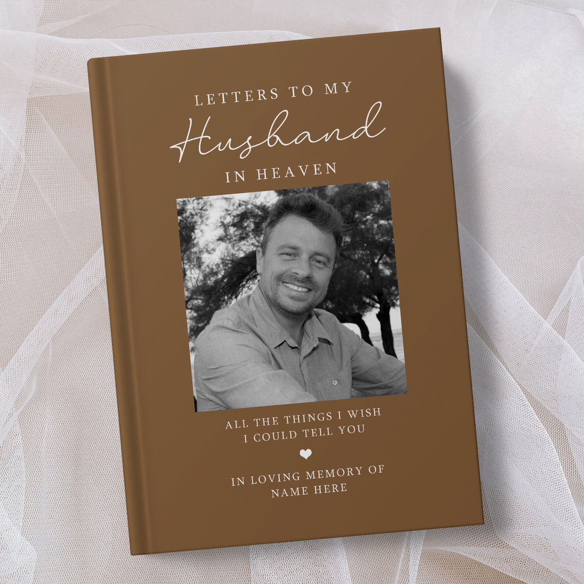 A personalised hardcover book titled 'Letters to My Husband in Heaven' with a brown cover, white elegant text, and a black and white photo of a smiling man. The book is displayed on a soft, draped white fabric background.