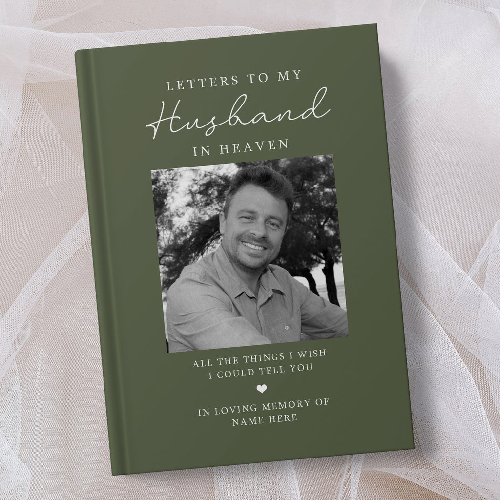 A personalised hardcover book titled 'Letters to My Husband in Heaven' in olive green, featuring a black and white photo of a smiling man and placeholder text, displayed on a soft beige fabric background.