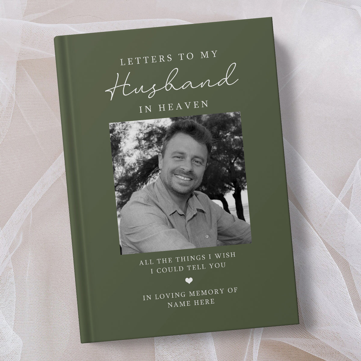 A personalised hardcover book titled 'Letters to My Husband in Heaven' in olive green, featuring a black and white photo of a smiling man and placeholder text, displayed on a soft beige fabric background.