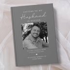 A personalised hardcover book titled 'Letters to My Husband in Heaven' with a grey cover, white script text, and a black and white photo of a smiling man. The book is photographed on soft white tulle fabric.