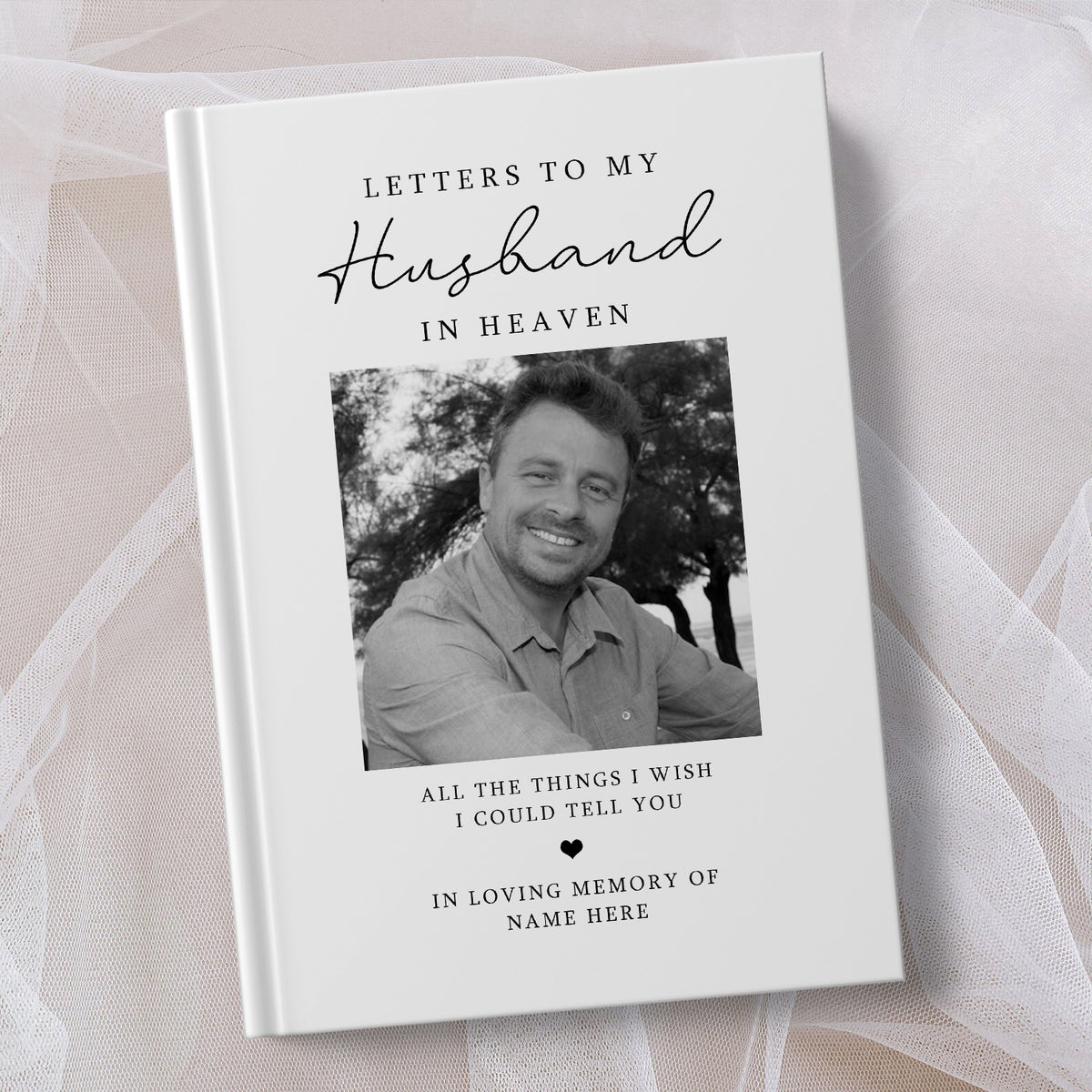 A personalised hardcover book titled 'Letters to My Husband in Heaven' with a black and white portrait photo of a smiling man on the cover, photographed on soft white tulle fabric.