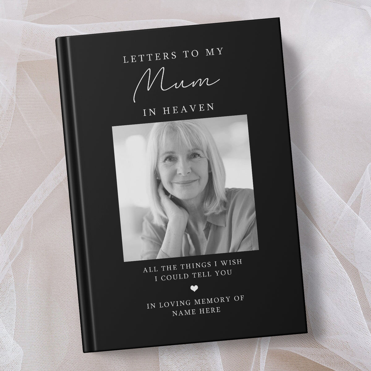A personalised hardcover book titled 'Letters to My Mum in Heaven' with a black cover, white text, and a black and white portrait photo of a woman. The book is displayed on a soft, draped white fabric background.