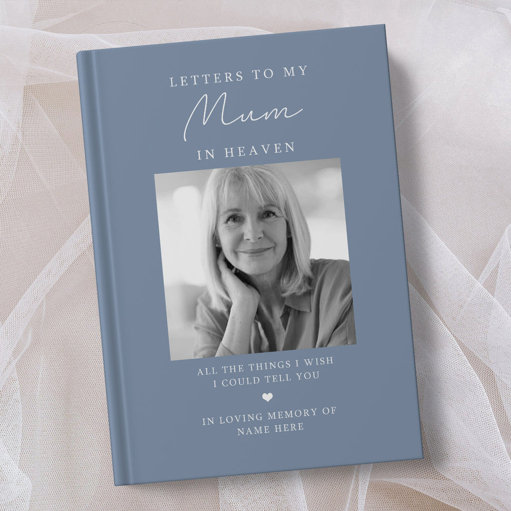 A personalsed hardcover book titled 'Letters to My Mum in Heaven' with a muted blue cover, featuring a black and white portrait photo of a woman and memorial text, displayed on soft white fabric.