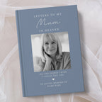A personalsed hardcover book titled 'Letters to My Mum in Heaven' with a muted blue cover, featuring a black and white portrait photo of a woman and memorial text, displayed on soft white fabric.