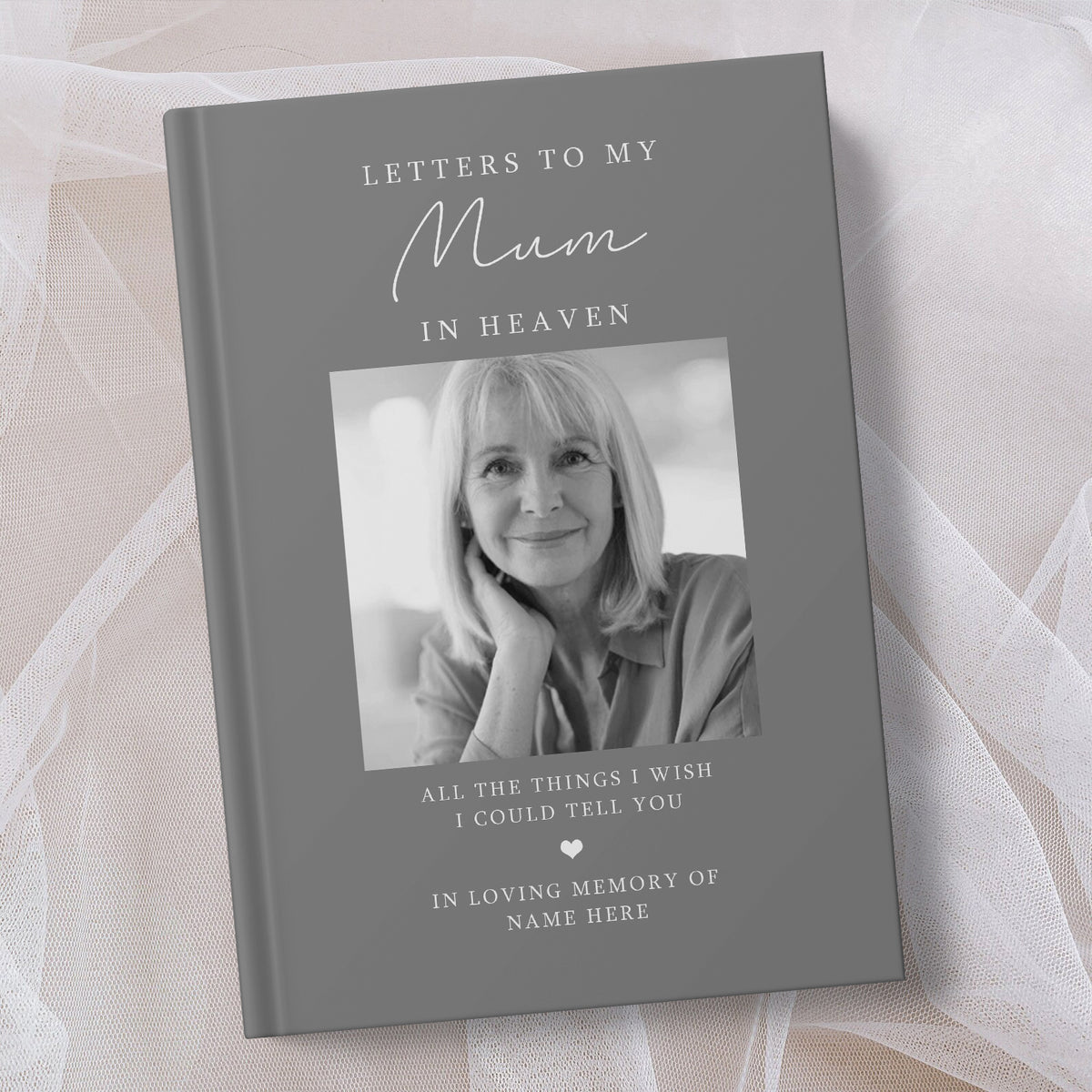 A personalised hardcover book titled 'Letters to My Mum in Heaven' with a grey cover, a black and white photo of a woman, and placeholder text for a memory message, displayed on soft white tulle fabric.