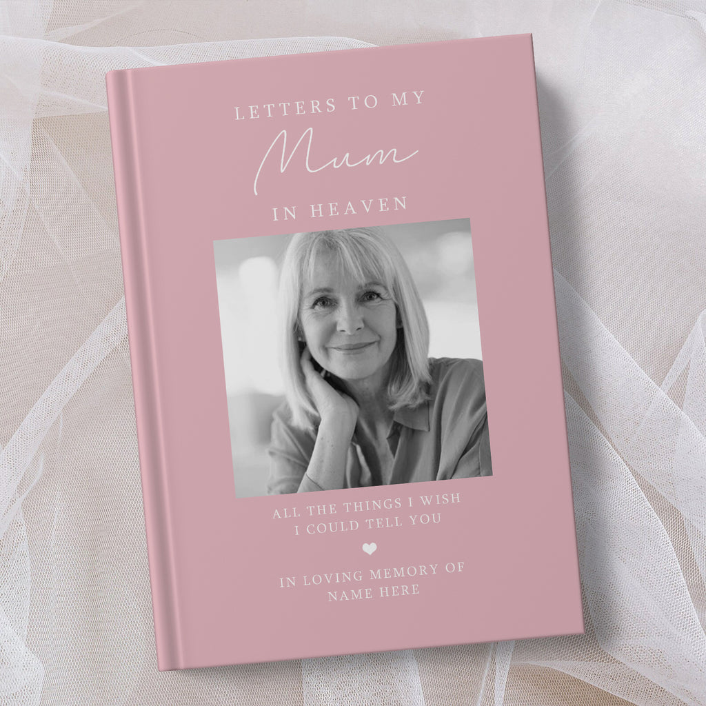A personalised hardcover book titled 'Letters to My Mum in Heaven' in dusty pink, featuring a black and white portrait photo of a woman on the cover, styled on a soft white fabric background.