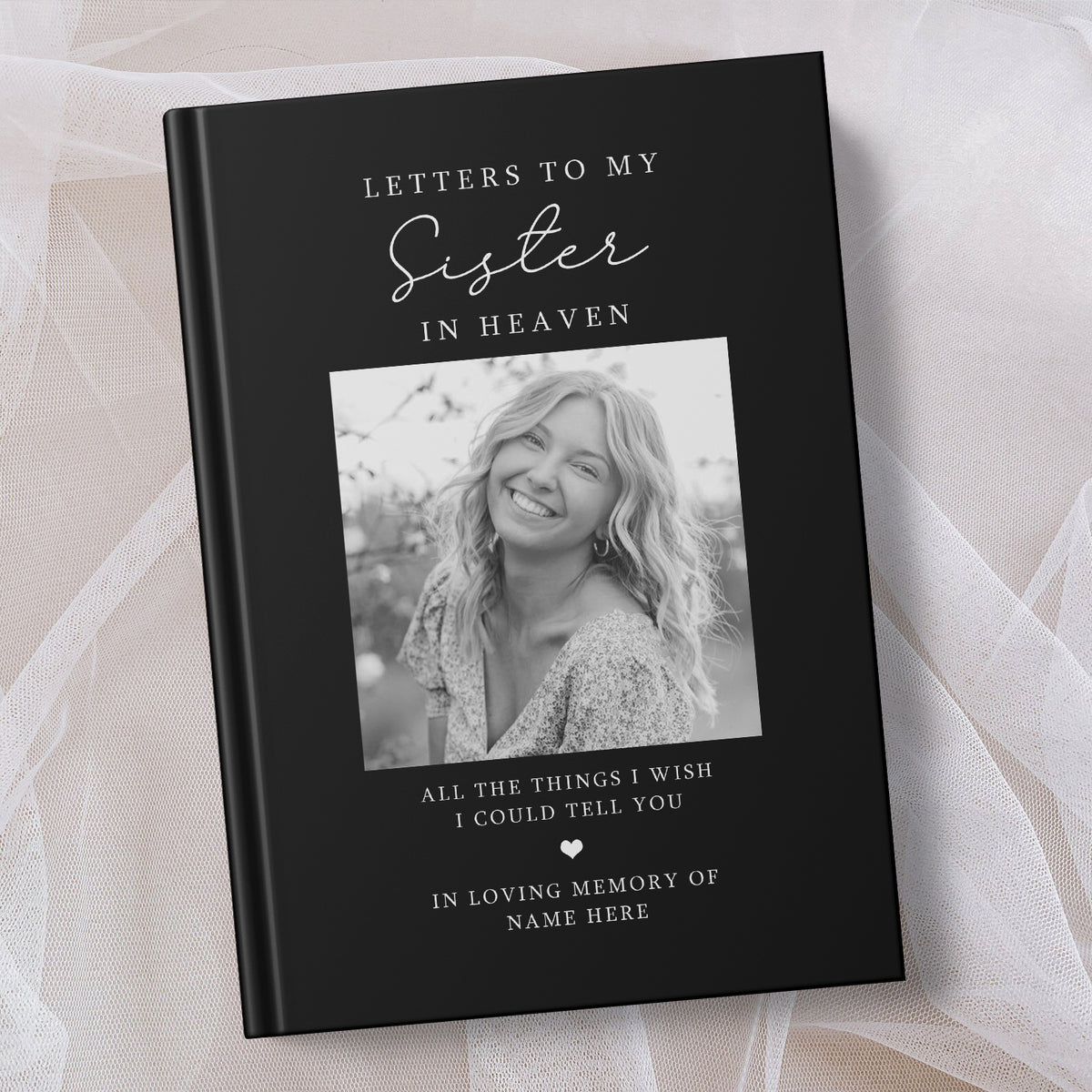 A black hardcover book titled 'Letters to My Sister in Heaven' with a black and white photo of a smiling woman on the cover, displayed on a soft beige fabric background.