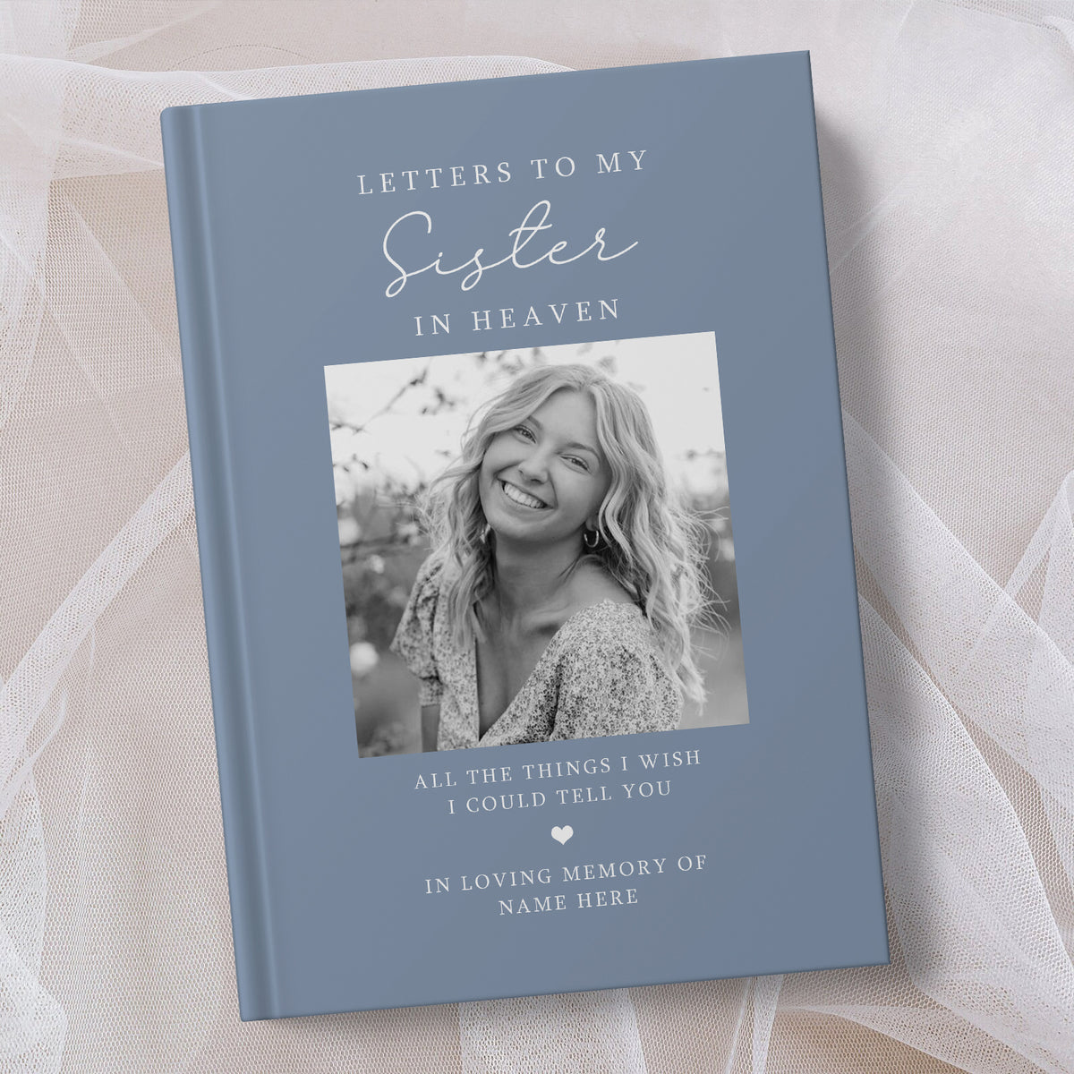 A personalised hardcover book titled 'Letters to My Sister in Heaven' with a dusty blue cover, featuring a placeholder photo of a smiling woman and customisable text, displayed on a soft beige fabric background.