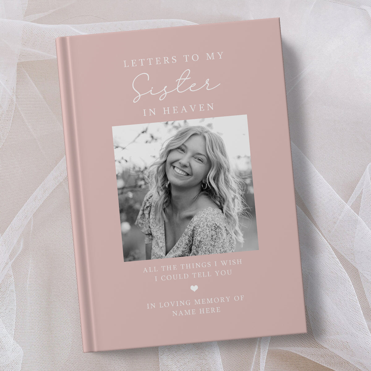 A personalised hardcover book titled 'Letters to My Sister in Heaven' with a dusty pink cover, featuring a black and white photo of a smiling woman and placeholder text, styled on a soft white fabric background.