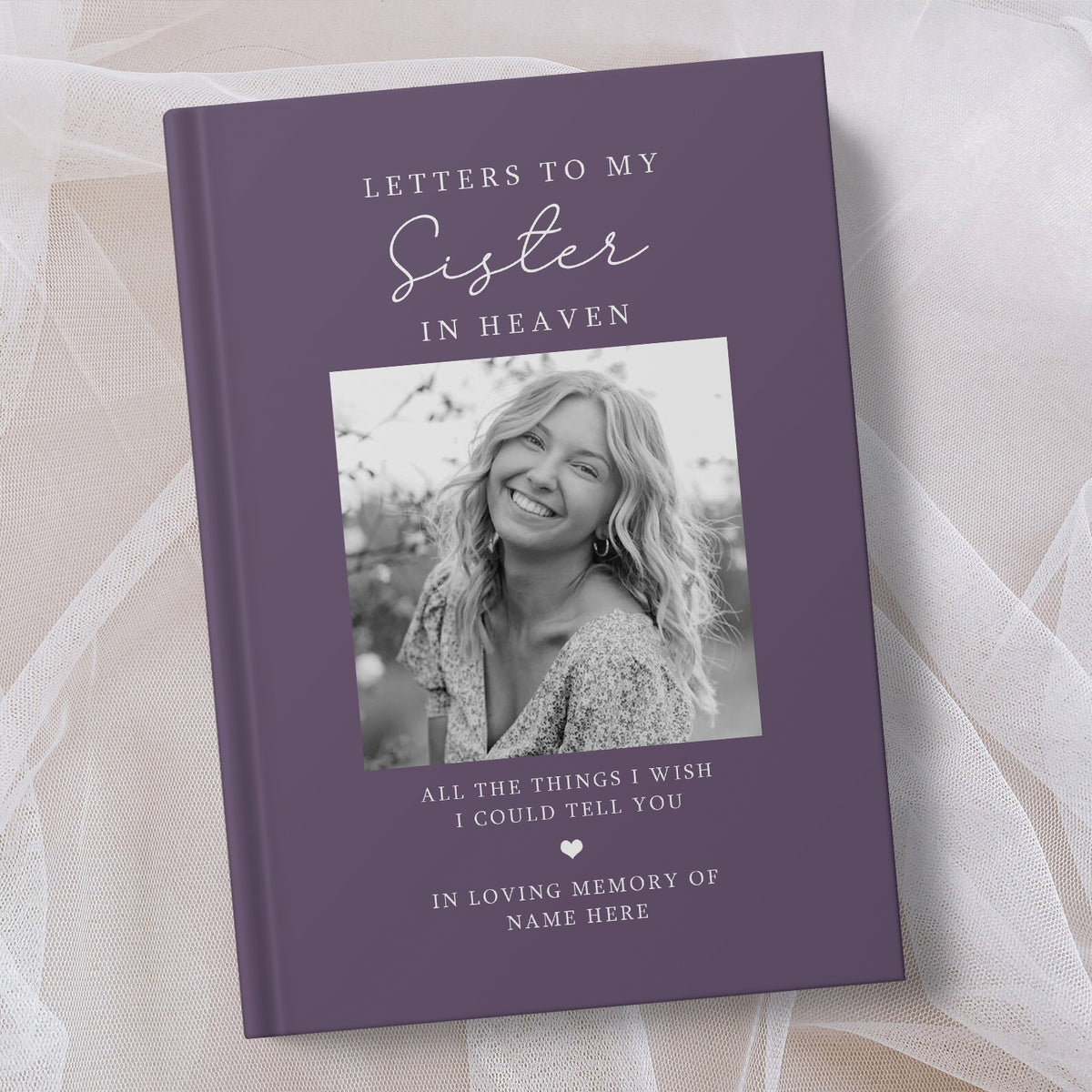 A personalised hardcover book titled 'Letters to My Sister in Heaven' in a muted purple colour, featuring a placeholder photo of a smiling woman on the cover. The book is displayed on a soft, draped white fabric background.
