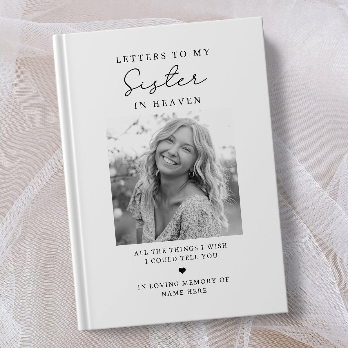A personalised hardcover book titled 'Letters to My Sister in Heaven' displayed on a soft, sheer white fabric background. The cover is white with elegant black typography and a central black and white photograph of a smiling woman.