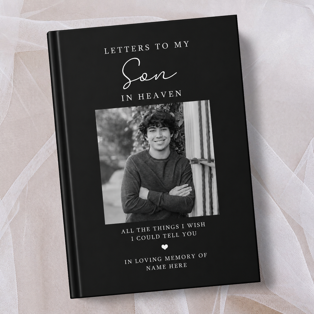 A black personalised hardcover book titled 'Letters to My Son in Heaven' with a placeholder photo of a young man, displayed on a soft beige fabric background.