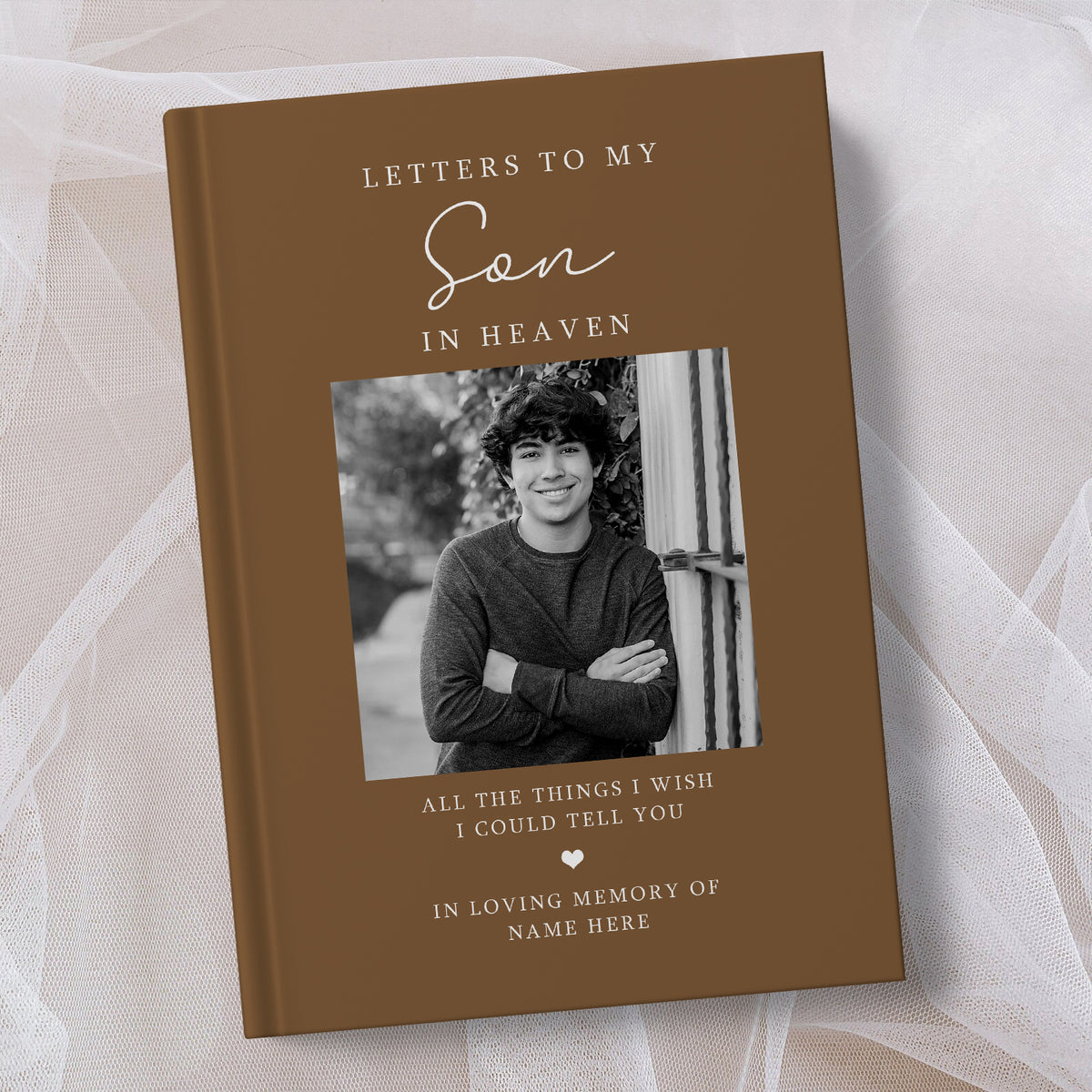 A personalised brown hardcover book titled 'Letters to My Son in Heaven' with a black and white photo of a young man on the cover, displayed on a soft white tulle fabric background.