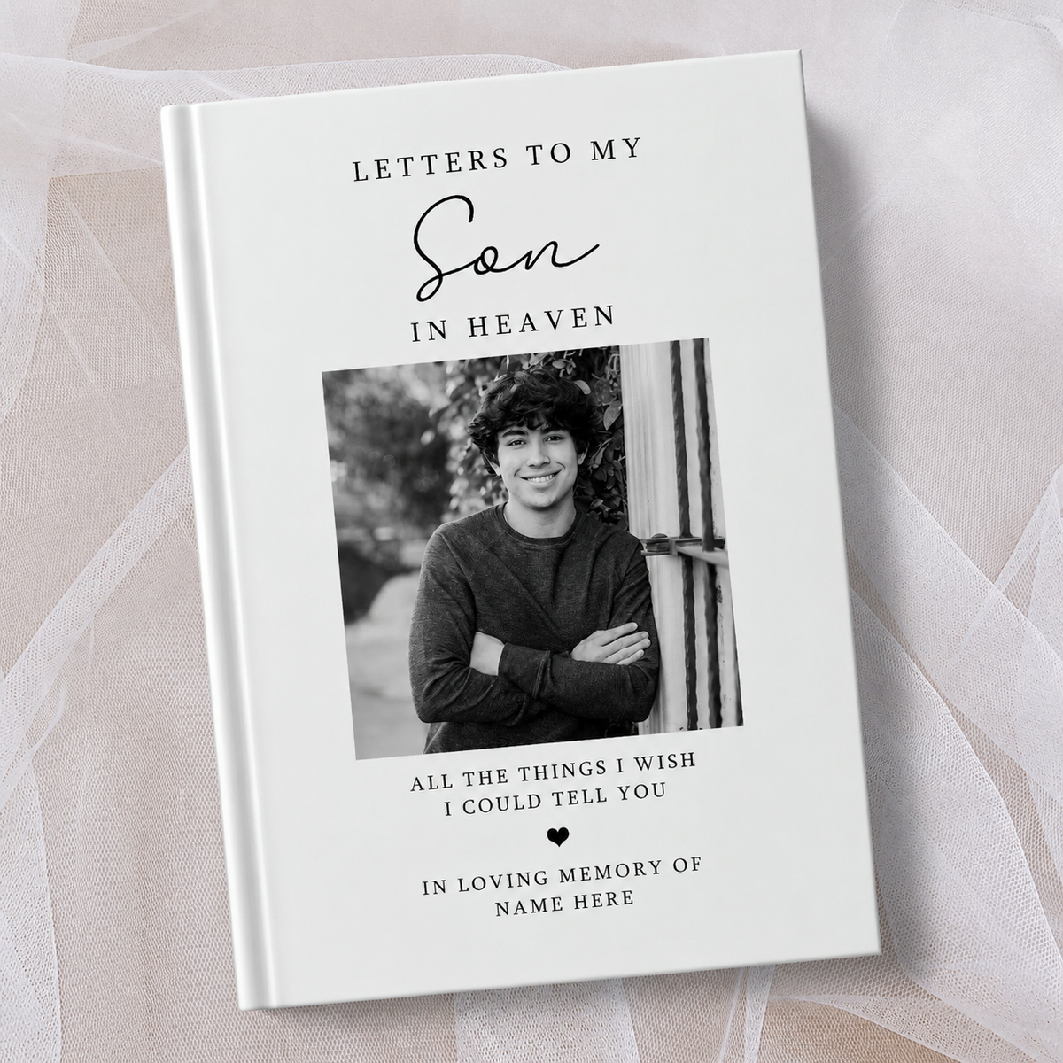 A personalised hardcover book titled 'Letters to My Son in Heaven' with a black and white photo of a young man on the cover, displayed on a soft beige fabric background.