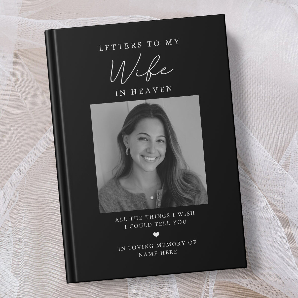 A personalised hardcover book titled 'Letters to My Wife in Heaven' with a black cover, white text, and a grayscale photo of a smiling woman, displayed on a soft beige fabric background.