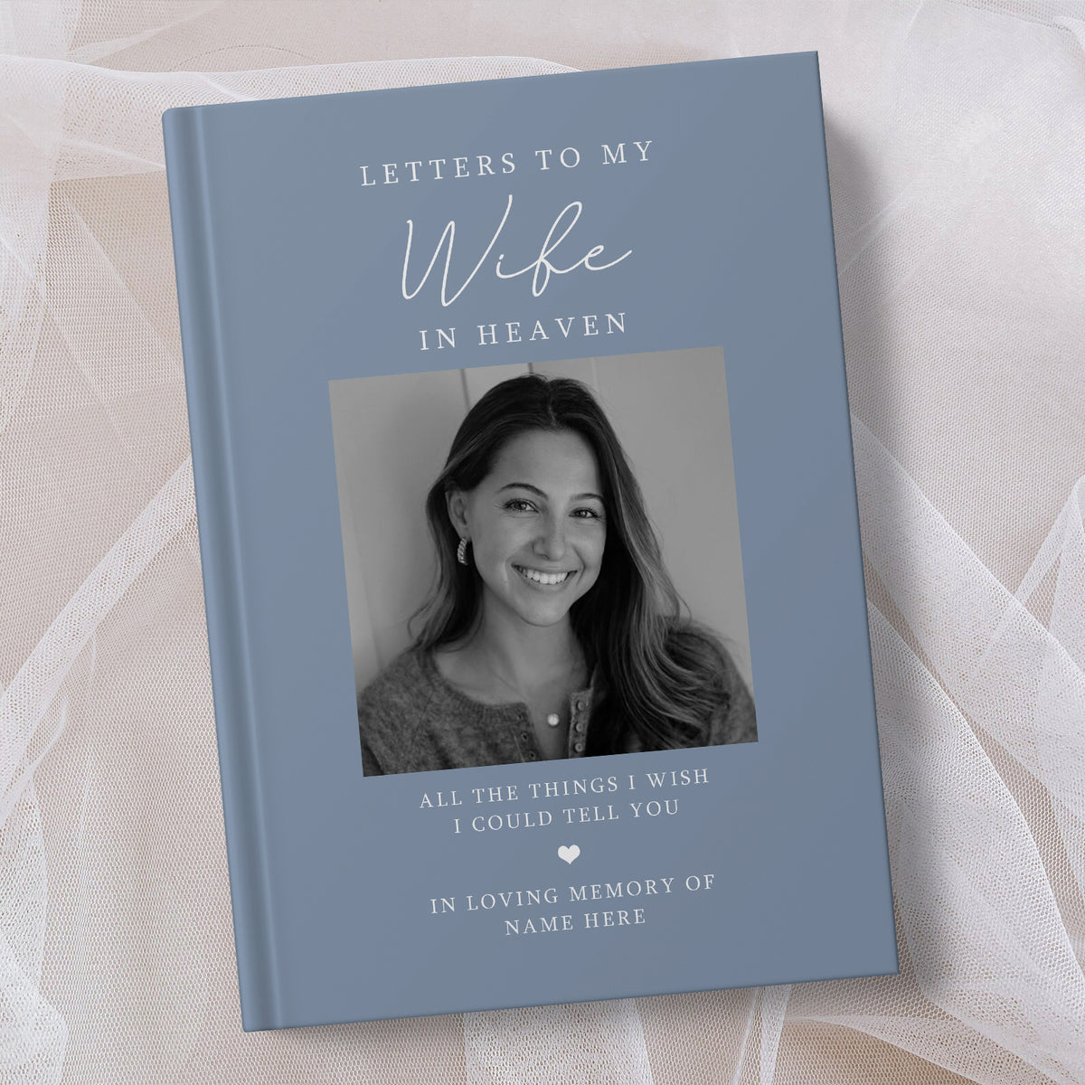 A personalised hardcover book titled 'Letters to My Wife in Heaven' with a dusty blue cover, featuring a black and white portrait of a smiling woman on the front. The book is displayed on a soft, draped white fabric background.