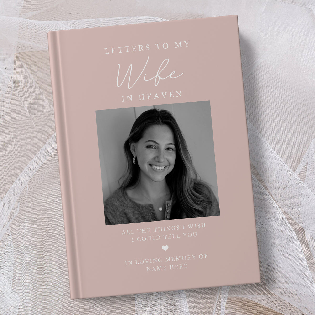 A personalised hardcover book titled 'Letters to My Wife in Heaven' with a dusty pink cover, featuring a black and white portrait photo of a smiling woman. The book is displayed on a soft, draped white fabric background.