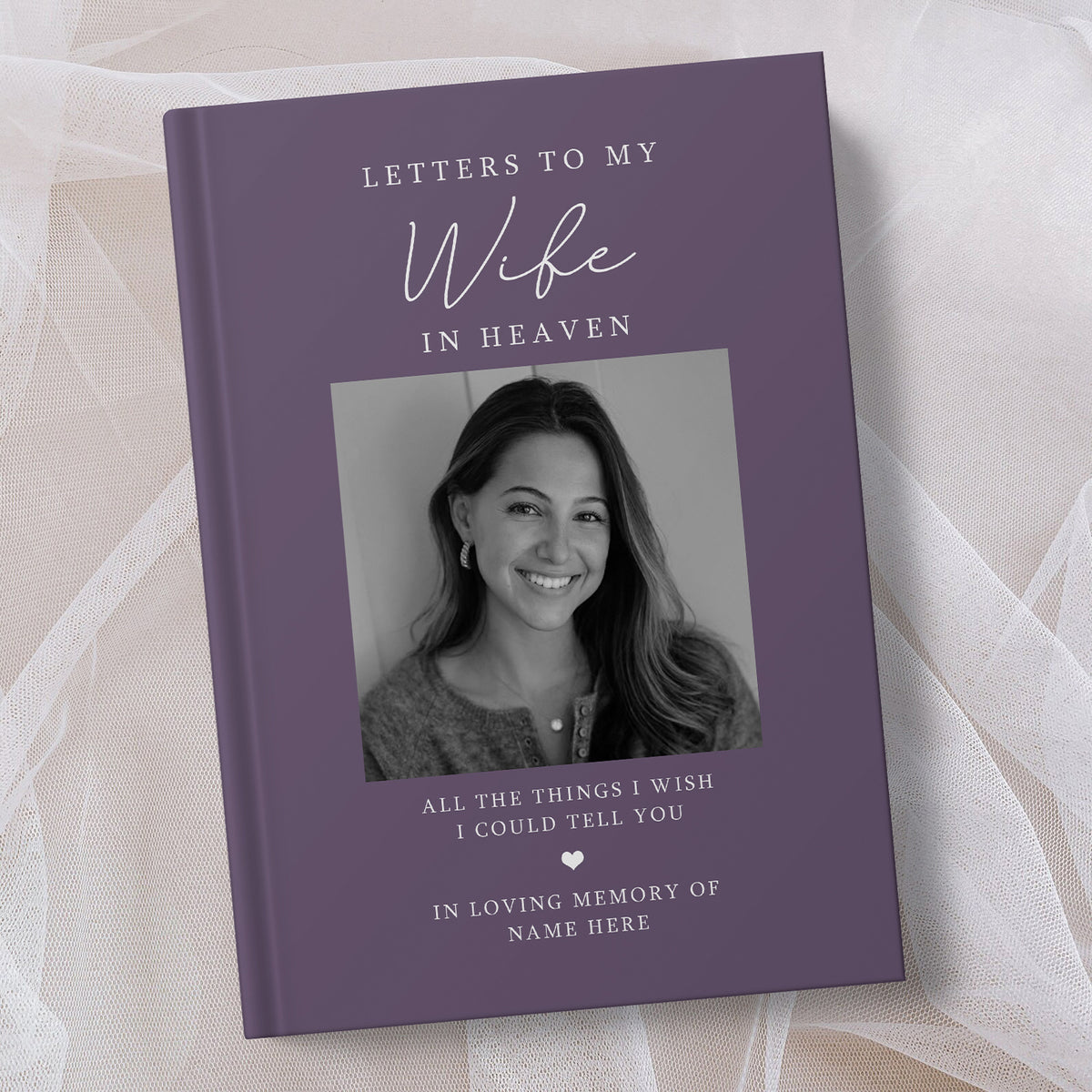 A personalised hardcover book titled 'Letters to My Wife in Heaven' with a purple cover, a black and white photo of a smiling woman, and memorial text, styled on a soft beige fabric background.