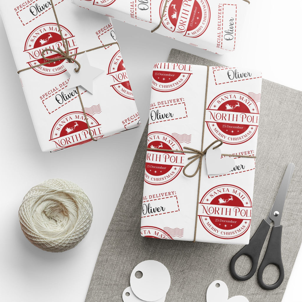 Three wrapped gift boxes tied with twine and decorated with matching white wrapping paper featuring red circular 'Santa Mail North Pole' stamps, arranged on a grey fabric surface with scissors, a spool of twine, and white gift tags on a white background.