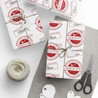 Three wrapped gift boxes tied with twine and decorated with matching white wrapping paper featuring red circular 'Santa Mail North Pole' stamps, arranged on a grey fabric surface with scissors, a spool of twine, and white gift tags on a white background.