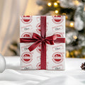 A gift box wrapped in white paper with a red circular 'North Pole' stamp pattern and a dark red satin ribbon tied in a bow, set against a softly blurred Christmas tree background.