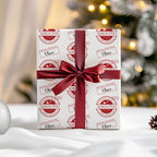 A gift box wrapped in white paper with a red circular 'North Pole' stamp pattern and a dark red satin ribbon tied in a bow, set against a softly blurred Christmas tree background.