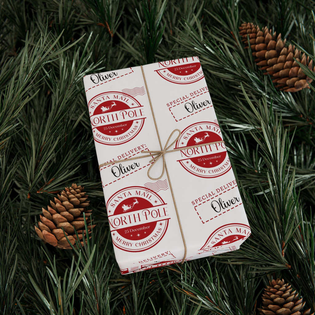 A wrapped gift box tied with twine, covered in repeating red and white North Pole Christmas stamp designs, placed on green pine branches with pinecones.