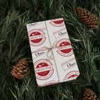 A wrapped gift box tied with twine, covered in repeating red and white North Pole Christmas stamp designs, placed on green pine branches with pinecones.
