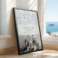 A framed personalised map print leaning against a window on a wooden floor, showing a faint map background, the text 'Our First Home' with names and date, and a black-and-white photo of a family at the bottom. A real interior setting and ocean view are visible through the window.